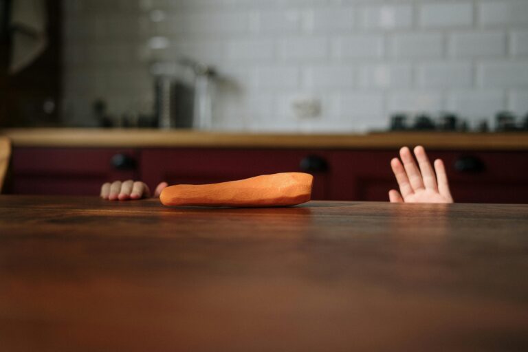 A child playfully reaching for a carrot on a wooden kitchen table indoors.